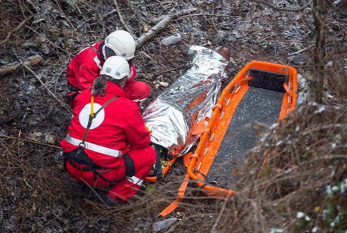 Two rescue workers in red uniforms assist a person wrapped in a thermal blanket on a stretcher in a wooded area.
