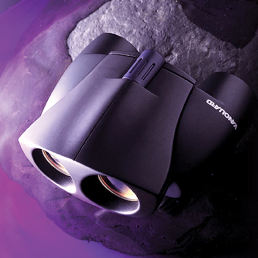A pair of black Vanguard binoculars rests on a dark, rock-like surface with a purple hue in the background.