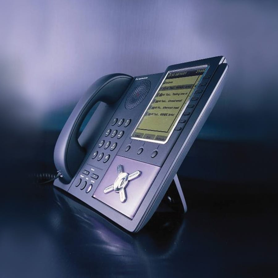 Office desk phone with a large digital display screen showing text, numerous buttons, and a corded receiver on a dark surface.